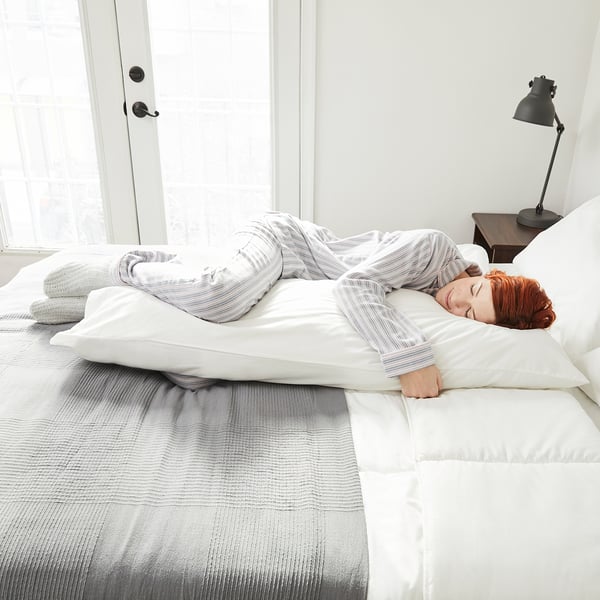 Person resting on side with long body pillow supporting the person back and knees. White bedding, striped pillow. Bedside lamp nearby.
