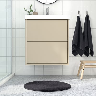 Modern bathroom with light tan BACKMÅRA mat, black towels, small wooden stool, grey round rug on speckled tile floor.
