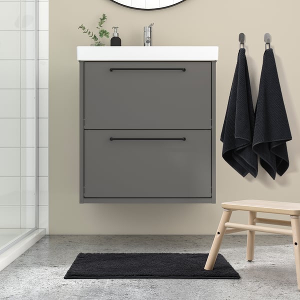 Modern bathroom with BACKMÅRA grey bath mat, grey vanity, wooden stool, dark towels, and glass shower door.