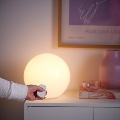 A person adjusts a spherical smart lamp on a white table with a remote control. The table also holds pink decor items and a poster.