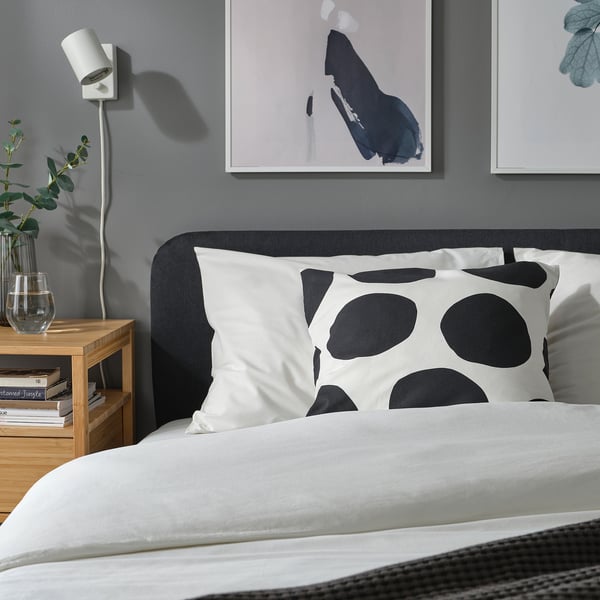 Minimalist black & white bedroom with large dotted pillow, bedside table greenery, books, abstract art, and wall light.