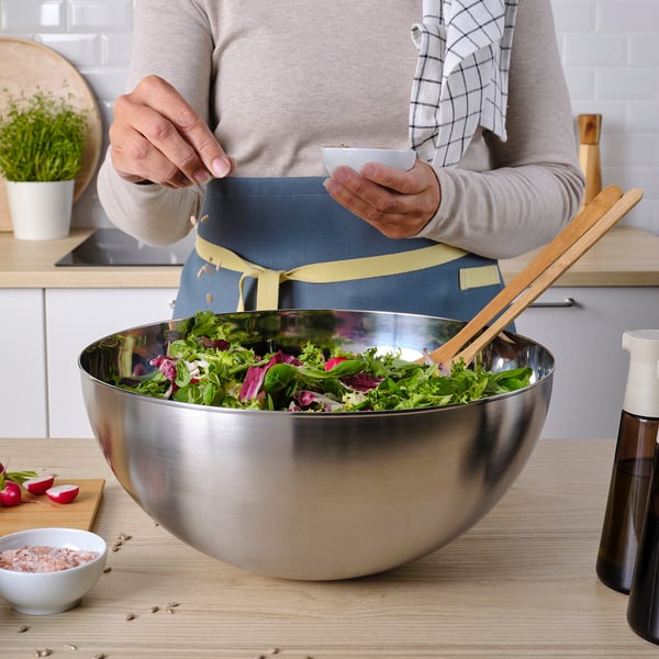 Person prepares salad in a large stainless steel bowl on a wooden countertop. The bowl has a modern, functional design.