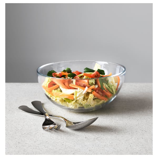 Clear glass bowl with salad, two metal serving spoons. Minimalist kitchen scene.