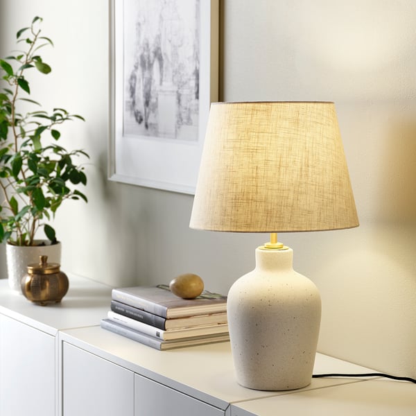 BLIDVÄDER lamp with beige linen shade & speckled base next to books, plant, and bowl, emitting warm light.