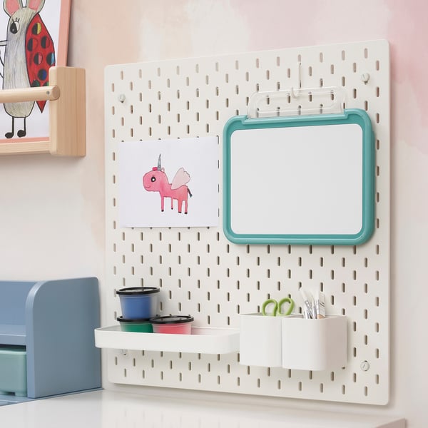 White wall-mounted pegboard with a teal-framed magnetic drawing board and containers holding coloured cups and scissors.