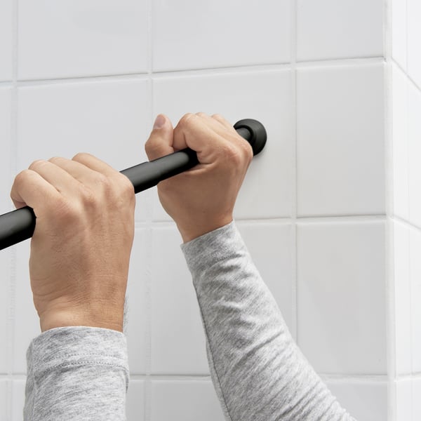 Person instals black shower curtain rod on tiled wall, using spring mechanism.