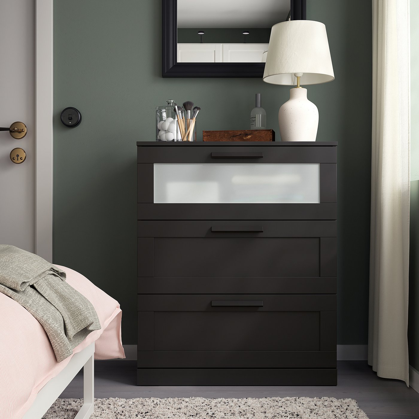 Black BRIMNES chest of drawers with frosted glass top, situated between a bed and a curtain, reflecting a stylish and organised bedroom setup.