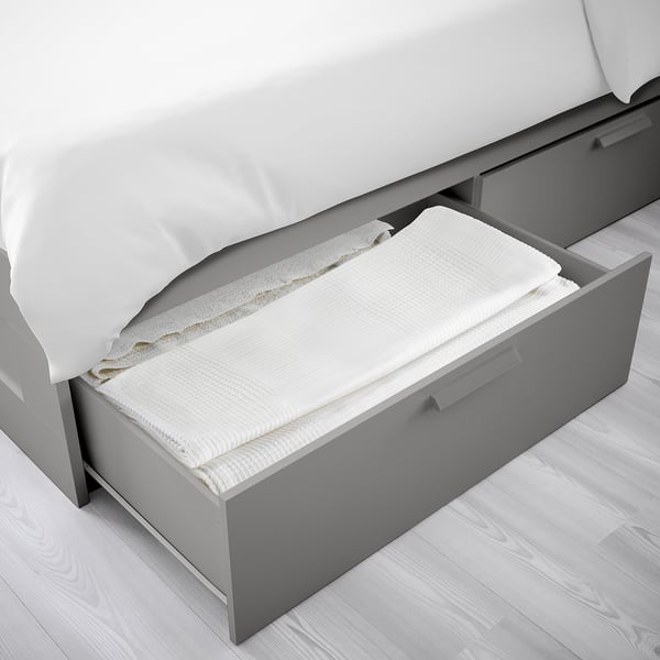 Grey bed frame with open storage drawers holding folded white linens.