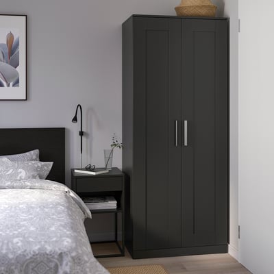 Modern bedroom with black BRIMNES wardrobe, grey comforter, bedside table, lamp, book, and plant.
