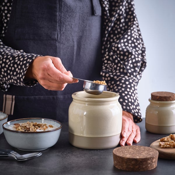 A person scoops granola from a rustic stoneware jar with a cork lid onto a bowl, demonstrating its use for food storage and serving.