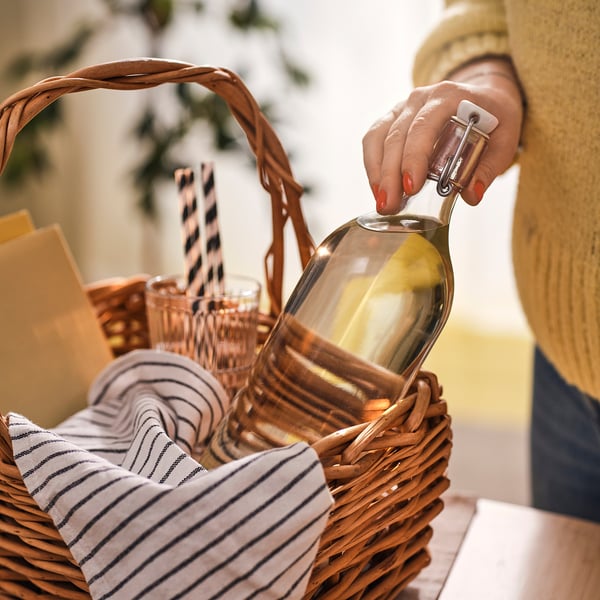 A person holding a wicker basket with a bottle of golden DRYCK FLÄDER, striped napkins, and glasses.