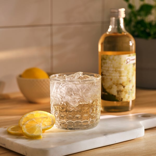 A glass of light, refreshing drink with ice and lemon. Behind it, a golden bottle with yellow flowers. In front, sliced lemons on marble.