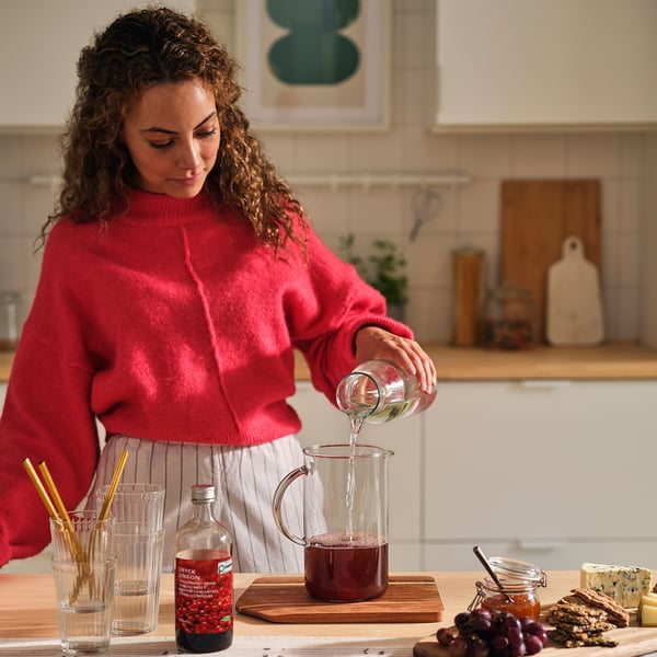 A person pours a dark red syrup into a pitcher on a kitchen counter, surrounded by glasses and ingredients.