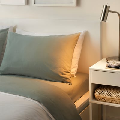 Minimalist bedroom: DVALA grey-green cotton sheets on large bed, neat bedside table with lamp & decor, warm lighting.