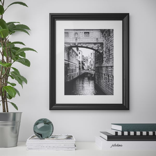 Black-framed a bridge over a canal hangs on a white wall. Below it, stacked books and a potted plant sit on a white surface.