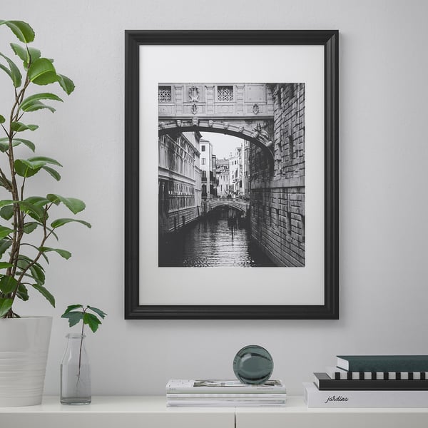 A black-framed black and white a bridge and canal hangs on a wall. On a shelf below, there are books, a plant, a vase, and a round object.