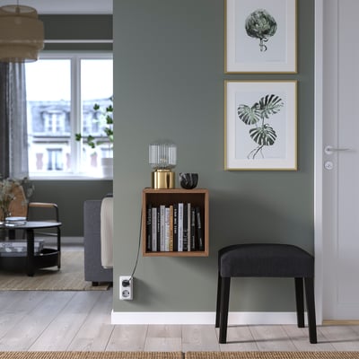 Wooden EKET wall cabinet with books, between art prints, grey stool below.