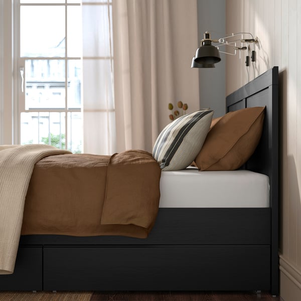 Modern bed frame with drawers and adjustable slats, tan bedding and pillows, black oak finish, wall-mounted reading light.