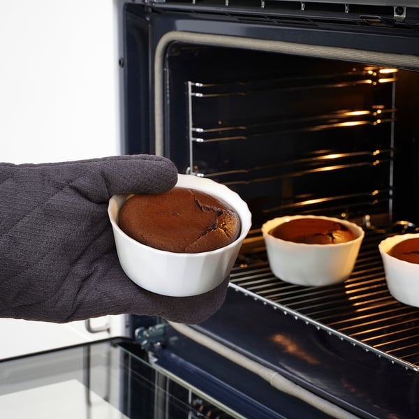 Person wearing an oven mitt holds a white, oven-safe dish with bake chocolate dessert. Inside the oven, similar dishes are placed on the rack.