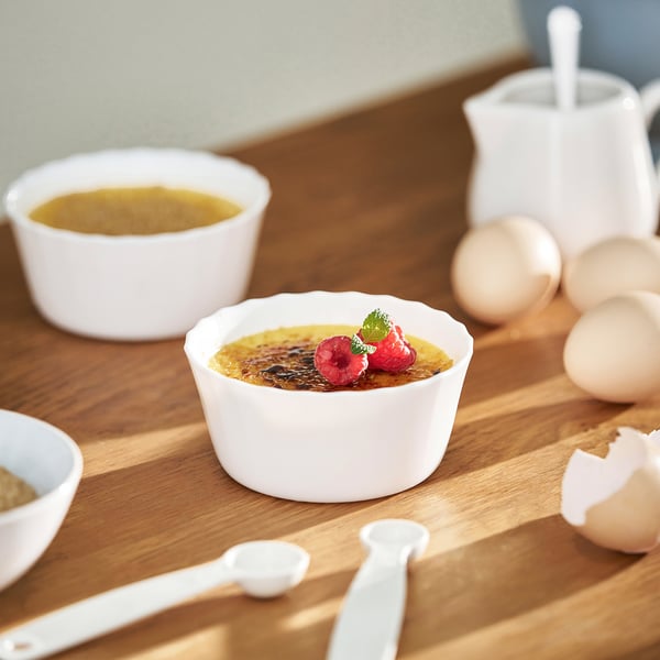 Ceramic ramekins on wooden table with crème brûlée, berries, mint, eggs, and utensils, suggesting baking.