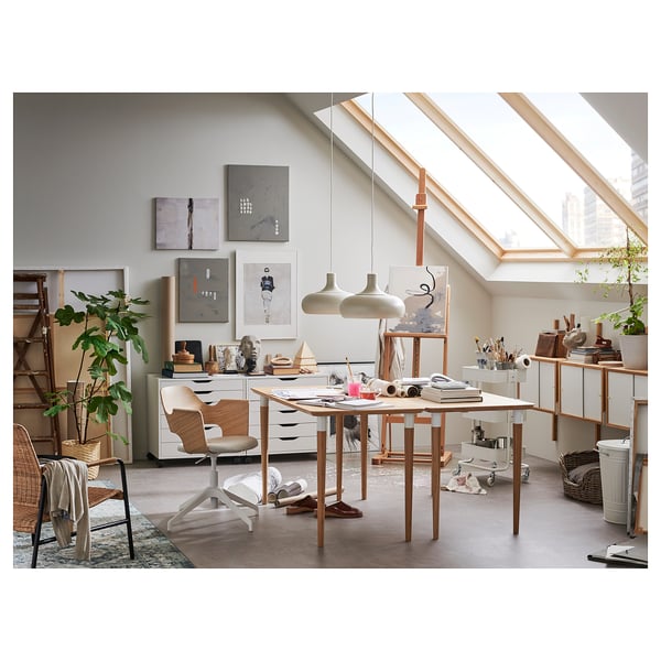 Bright, modern art studio with large skylights, wooden desk, chair, painting supplies, and various artworks on the wall.