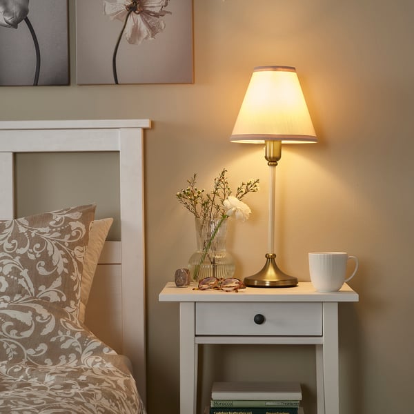 Bedside with FLYGHÖJD lamp. Cream lamp on white stand. Small vase with white flowers.