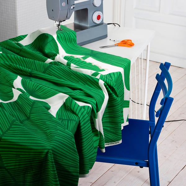 A sewing machine on a white table holds green and white leafy fabric. Blue chair, scissors, and thread nearby.