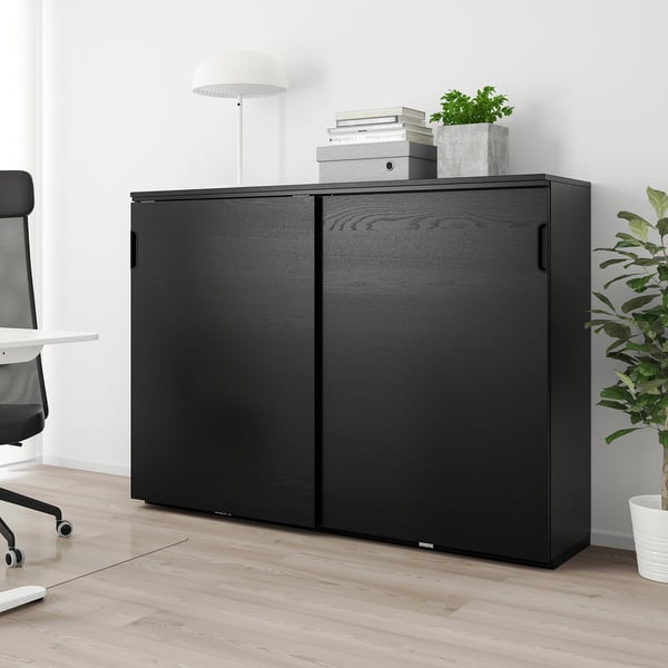 GALANT black cabinet, slim, tall, with sliding doors, in office setting with plants & chair.