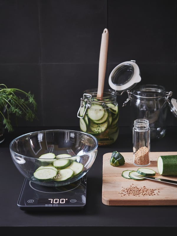 Digital kitchen scale displays 700g, holding glass bowl with sliced cucumbers. Nearby, jars, spices, chopping board with veggies and herbs.