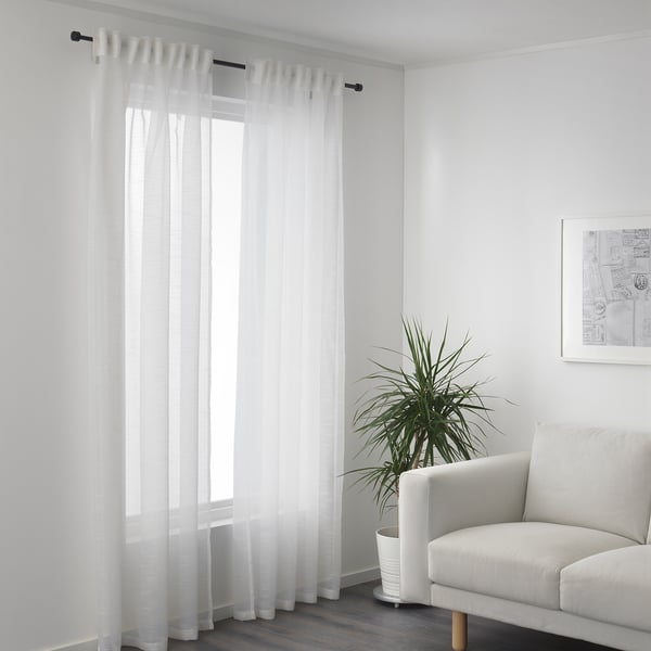 White curtains with sheer fabric hang by a window, allowing daylight while providing privacy. A plant and a white sofa are placed nearby.