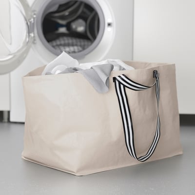Beige GÖRSNYGG laundry bag with striped handles, upright next to washing machine, filled with light clothes.