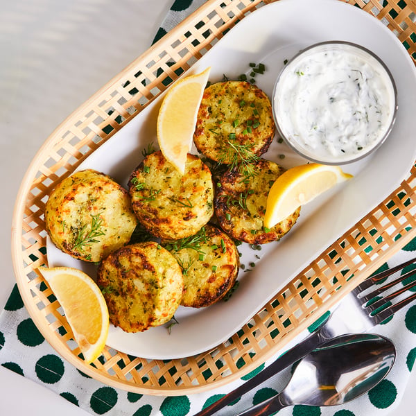 Golden-brown vegetable cakes served on a white plate with lemon wedges and a cup of sauce.