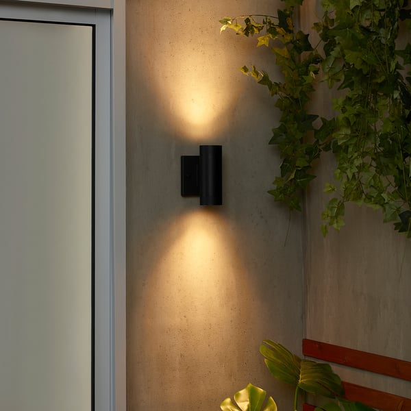 Black wall lamp GRÖNSPRÖT on an outdoor wall, emitting warm light, surrounded by plants. Appears compact compared to surrounding wall.