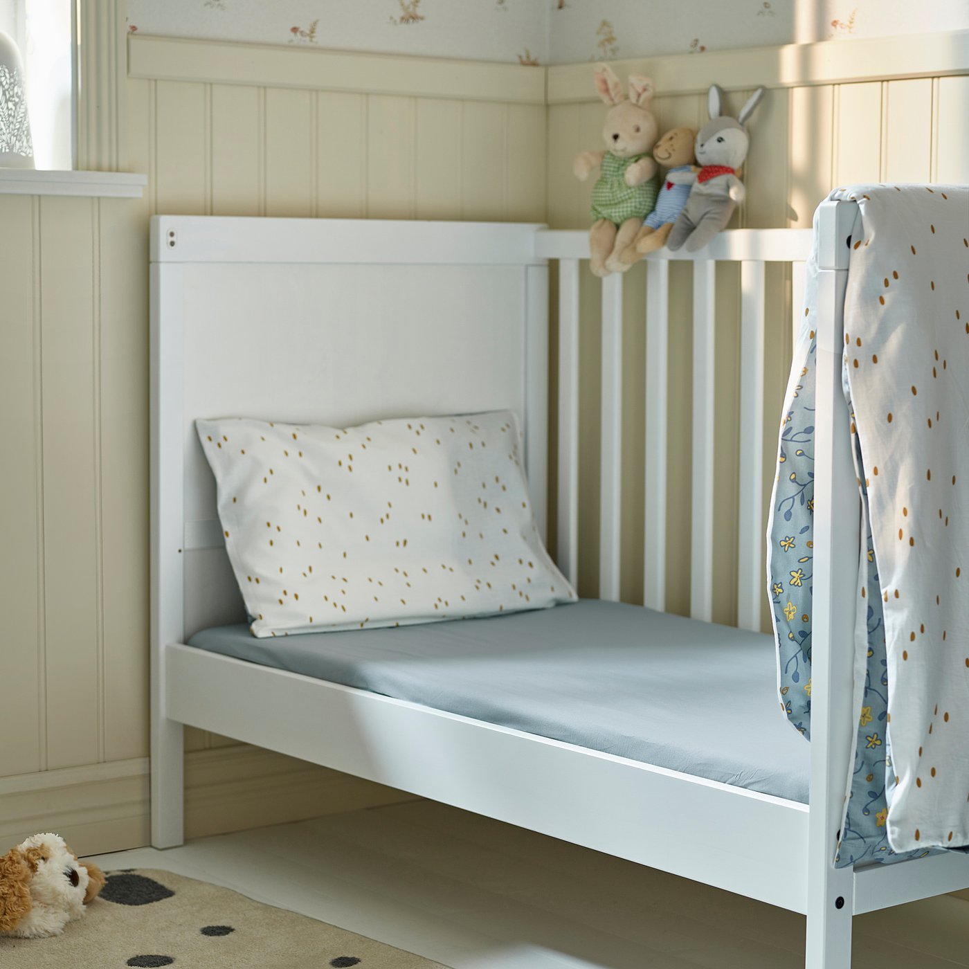 Small white GULDVÄVARE crib with blue mattress, soft toys, pattern blanket.