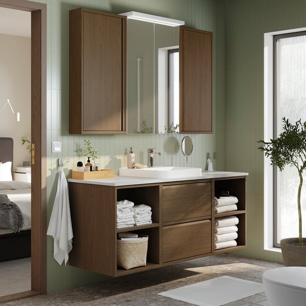 Modern bathroom with brown cabinet, open shelves, white towels, green tiles, and mirror with lights.
