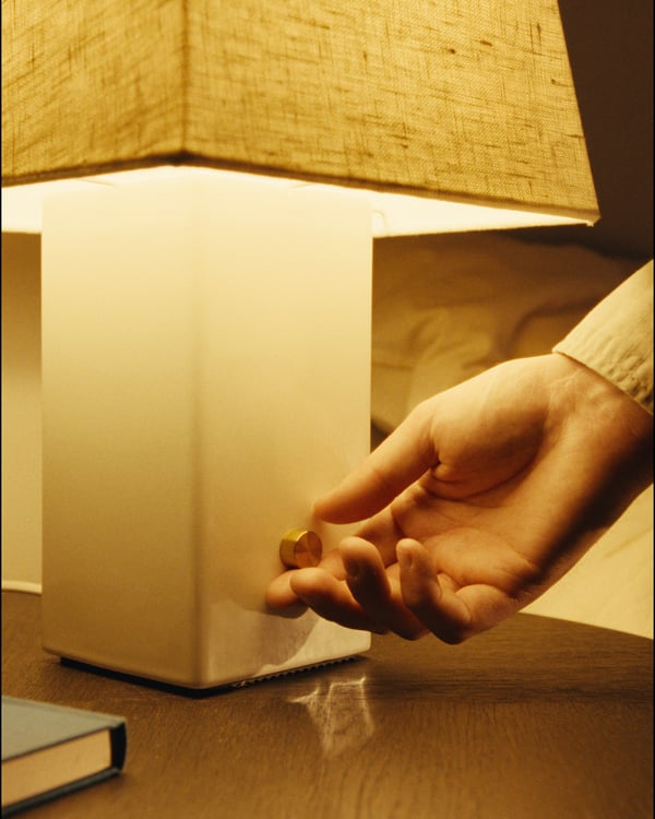 A hand adjusts a golden brass knob on a HALGATT table lamp. The lamp has a white ceramic base and a beige linen shade, casting warm light.