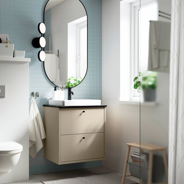Modern beige bathroom cabinet with blue-tiled walls. Round mirror, black fixtures, and plants.
