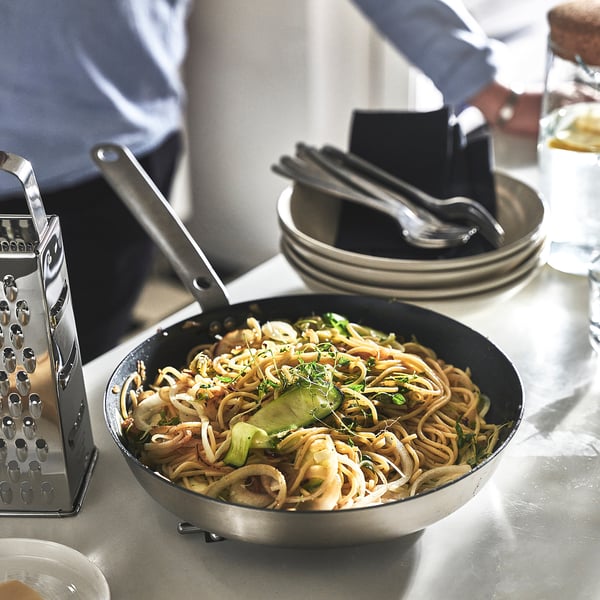 Cooking noodles and veggies. Tongs over pan, cheese grater, plates ready.