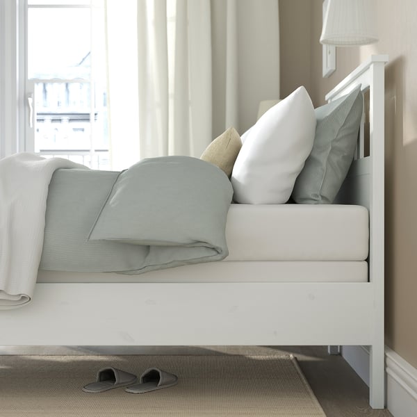 Cosy bed with blue blanket, pillows, and window curtains. Lamp and slippers nearby.