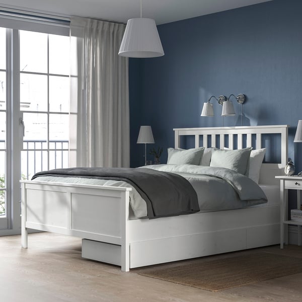 Small white wooden HEMNES bed with grey linens against a blue wall, featuring a large window and draped curtains.
