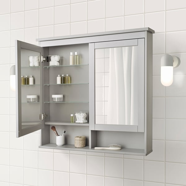Grey mirrored bathroom cabinet with multiple shelves displaying toiletries. Cabinet is mounted on tiled wall with lights on either side.