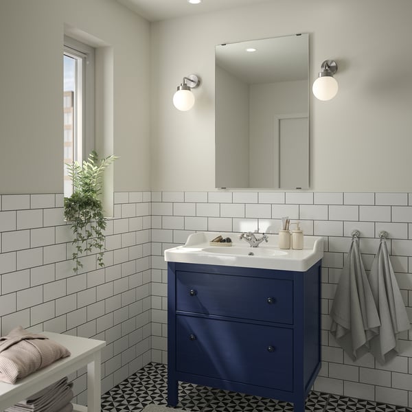 Modern bathroom with blue HEMNES cabinet, white sink, mirror, lights. Grey towels, green plant, patterned floor.