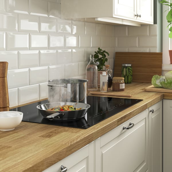 Modern kitchen with wooden counter, white tiles, black induction hob, stainless pan, colourful veggies, jars, cutting board, white bowl.