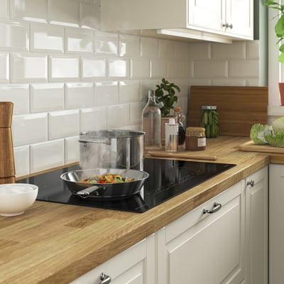 Modern kitchen with wooden counter, white tiles, black induction hob, stainless pan, colourful veggies, jars, cutting board, white bowl.