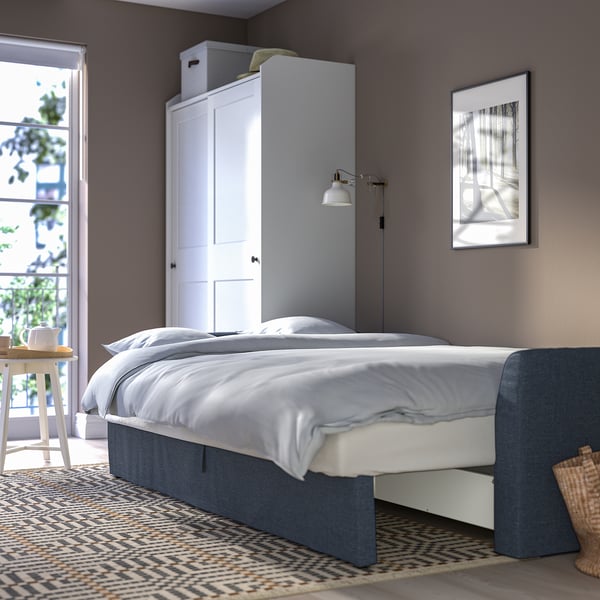 Modern bedroom with grey bed, white linens, blue headboard, trendy rug, side table with plants, and wall art. Tall white wardrobe behind bed.