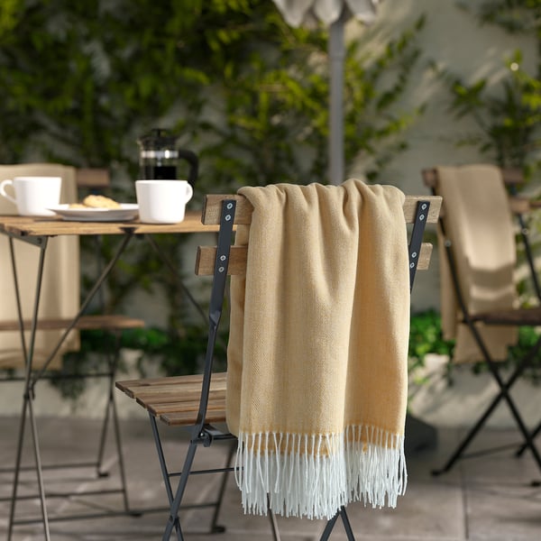 HOLMVI beige-yellow throw with fringes folded on chair, larger than coffee cups and plates on table.