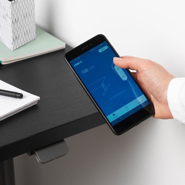 Person adjusts desk height using smartphone app. Smart desk app visible on phone screen.