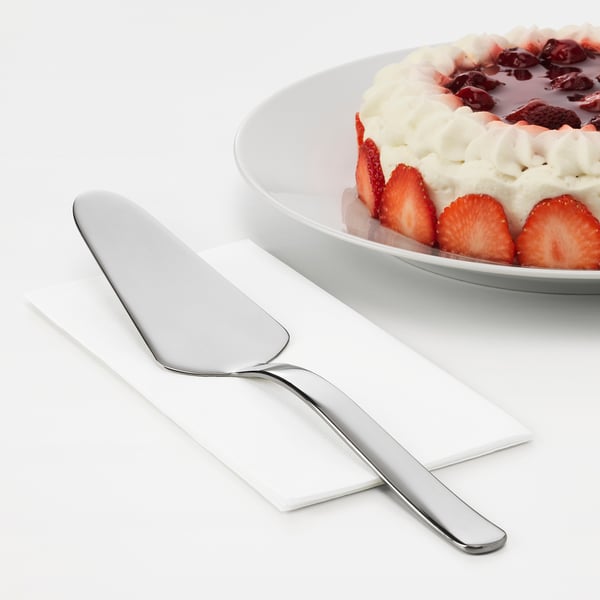 A sleek stainless steel cake slicer from IKEA 365+ series lies on a napkin, ready to serve a strawberry-topped cake on a white plate.