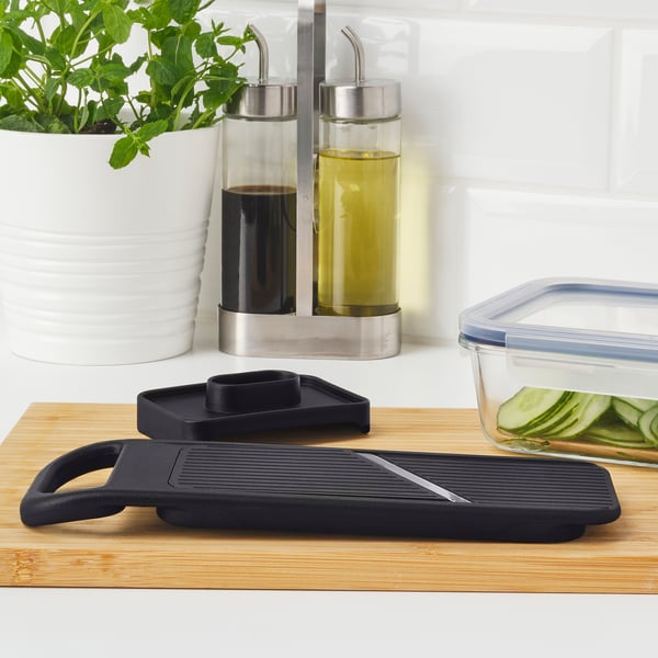 Black mandoline slicer on wooden board, with adjustable thickness dial and handguard, surrounded by glass containers and herbs.