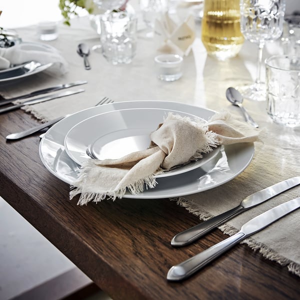 Table set with white porcelain plates, cutlery, and glasses. Placed on a wooden table with beige cloth napkins.